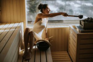Woman poring water on sauna rocks with ladle in sauna